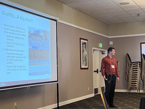 A man looks towards the audience while standing beside a projector screen with a slide title 'Graffiti...A big deal?'.